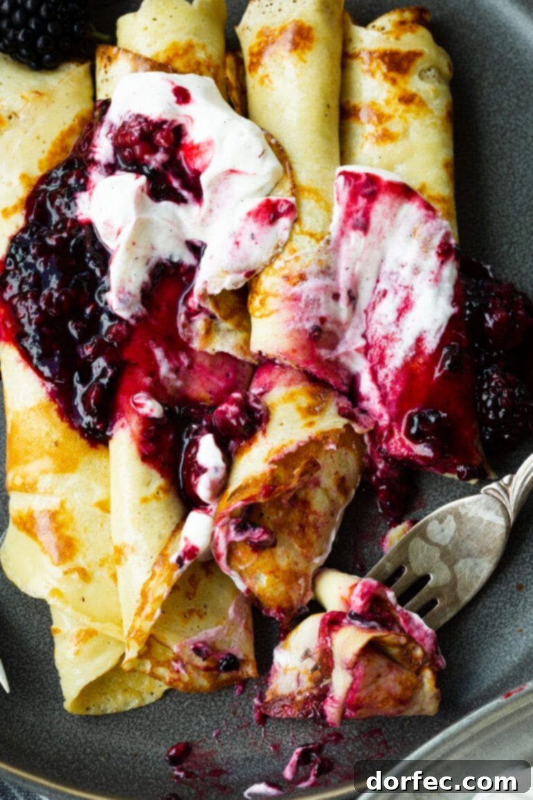 a photo of several rolled up finnish pancakes topped with blackberries and whipped cream and a fork cutting off a bite.