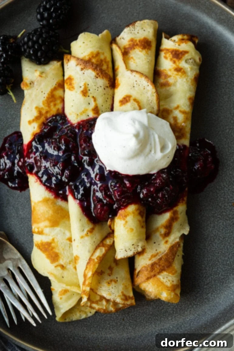a photo of several golden pan fried thin finnish pancakes that are rolled up and topped with a blackberry sauce and whipped cream