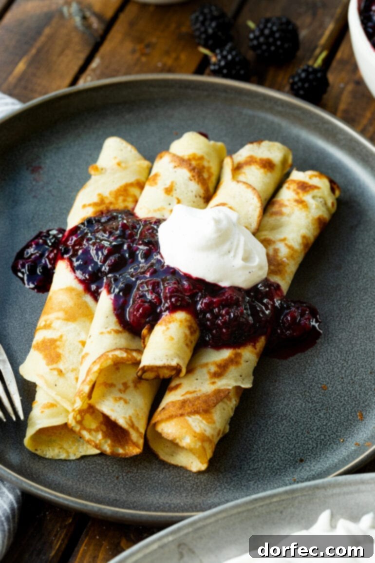 a photo of rolled up golden finnish pancakes topped with a thick blackberry sauce and a dollop of whipped cream.