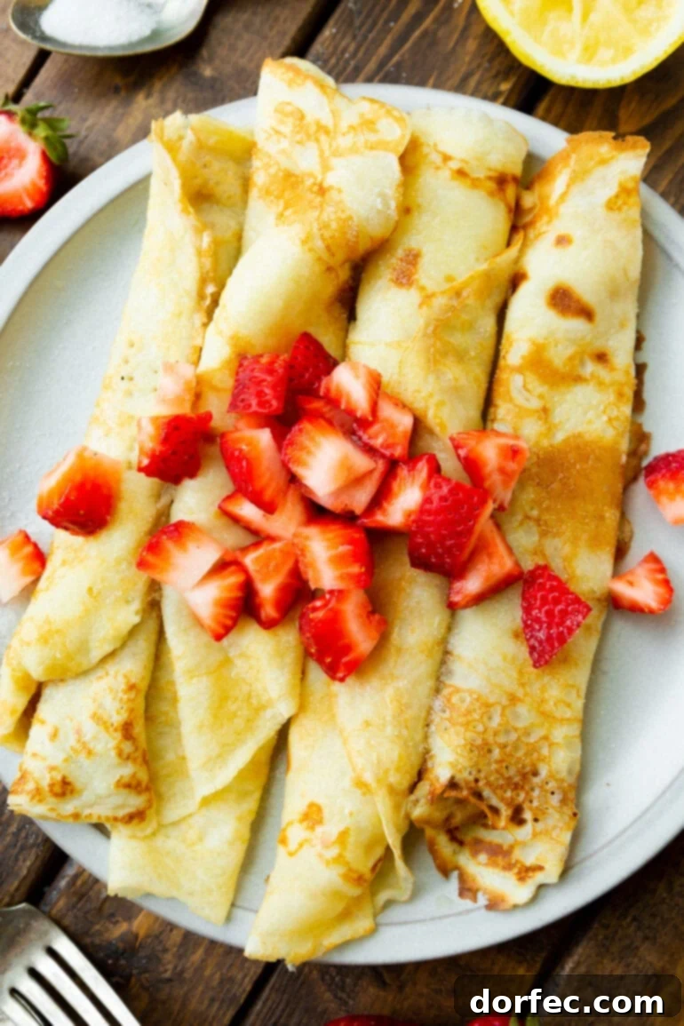 A beautiful photo of four golden Swedish pancakes, delicately rolled and arranged on a pristine white plate, garnished with vibrant diced strawberries. A perfect blend of simple elegance and irresistible flavor, ready to be enjoyed.