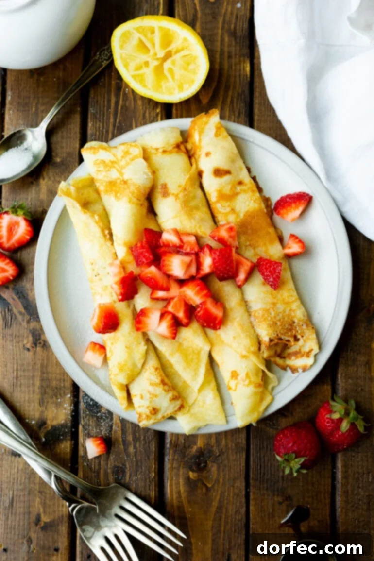 A close-up shot of four golden-brown Swedish pancakes, gracefully rolled up and presented side-by-side on a white plate. Freshly diced strawberries are scattered over the top, adding a pop of color and sweetness.