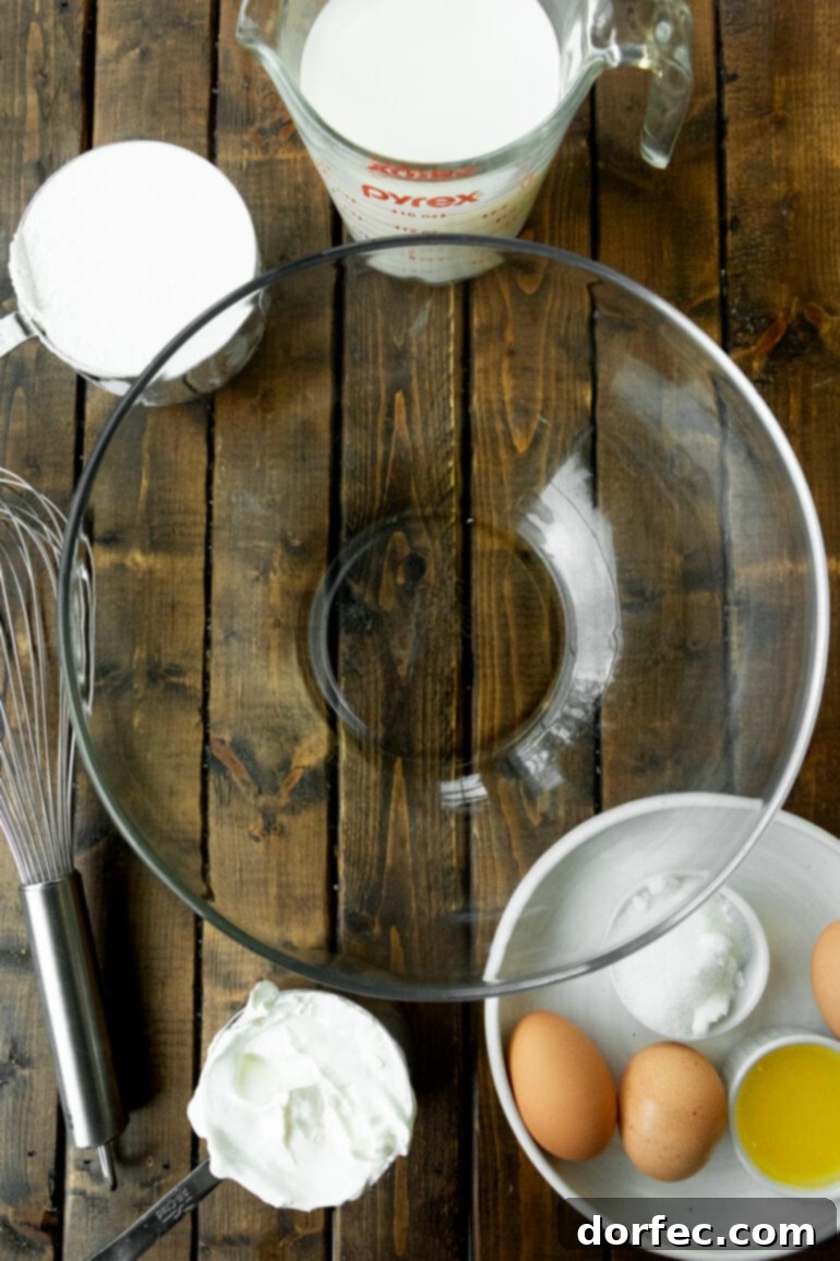 A large, empty glass mixing bowl sits prominently in the center of a clean white counter, surrounded by smaller bowls neatly arranged in a semicircle. Each smaller bowl holds a key ingredient for Swedish pancakes: flour, sugar, eggs, and melted butter, all ready for mixing. This setup suggests a well-prepared and easy-to-follow recipe, inviting home cooks to gather their pantry-friendly essentials.