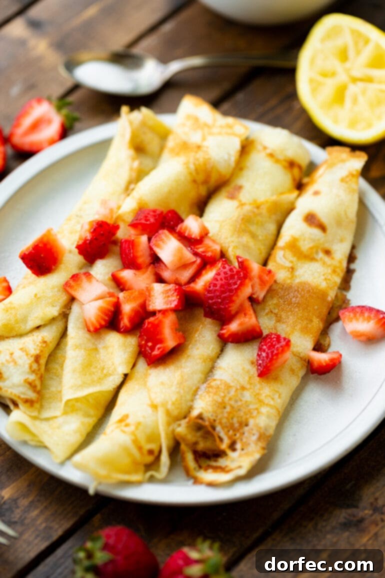 A stunning close-up of four perfectly rolled Swedish pancakes, each delicately golden and arranged side-by-side on a white plate. They are generously adorned with vibrant diced strawberries, presenting an inviting and elegant breakfast or dessert.