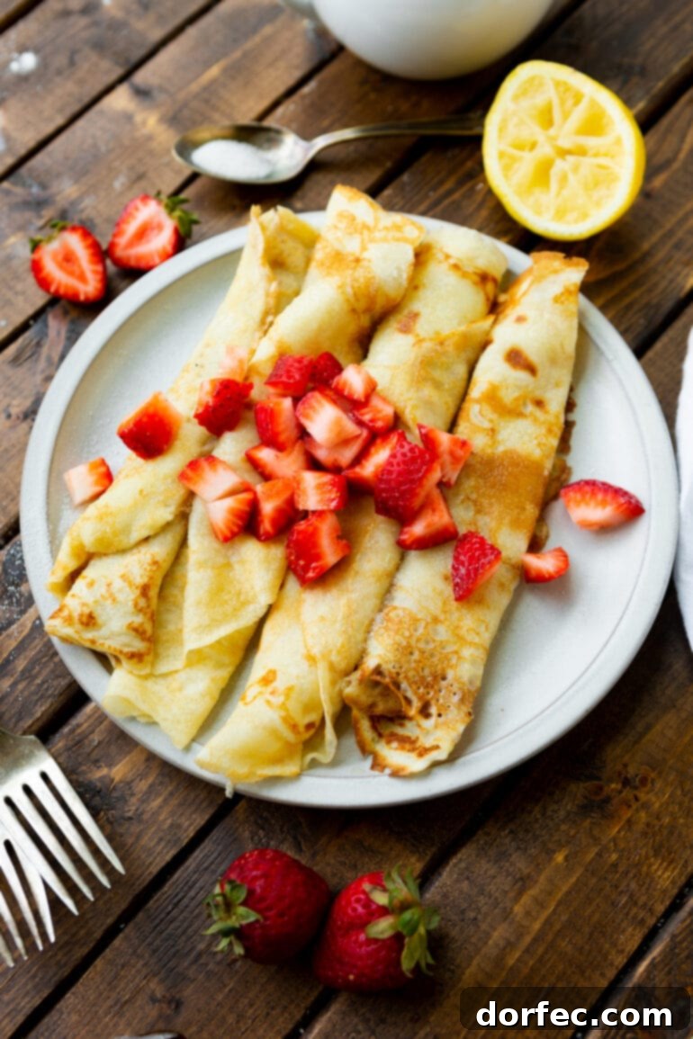 A crisp, high-angle shot reveals a white plate holding four perfectly rolled, golden-brown Swedish pancakes. Each one is generously topped with a sprinkle of bright red diced strawberries, and a silver fork rests casually beside the plate, inviting you to indulge in this delightful breakfast. The presentation is simple yet elegant, promising a delicious start to the day.