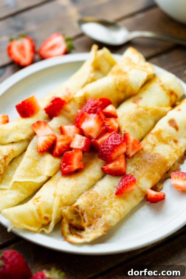 A delightful array of several golden-brown Swedish pancakes, beautifully rolled and nestled side-by-side on a crisp white plate. A generous scattering of bright red diced strawberries adorns the top, making for an inviting and elegant breakfast spread. The warmth of the pancakes and the freshness of the fruit promise a truly satisfying start to the day.