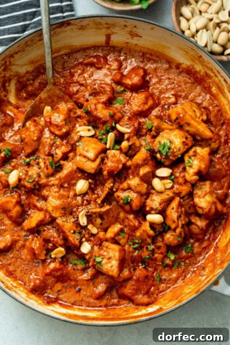 A photo of a large skillet filled with chicken chunks simmering in a thick, rich peanut butter sauce, garnished with fresh cilantro and chopped peanuts.