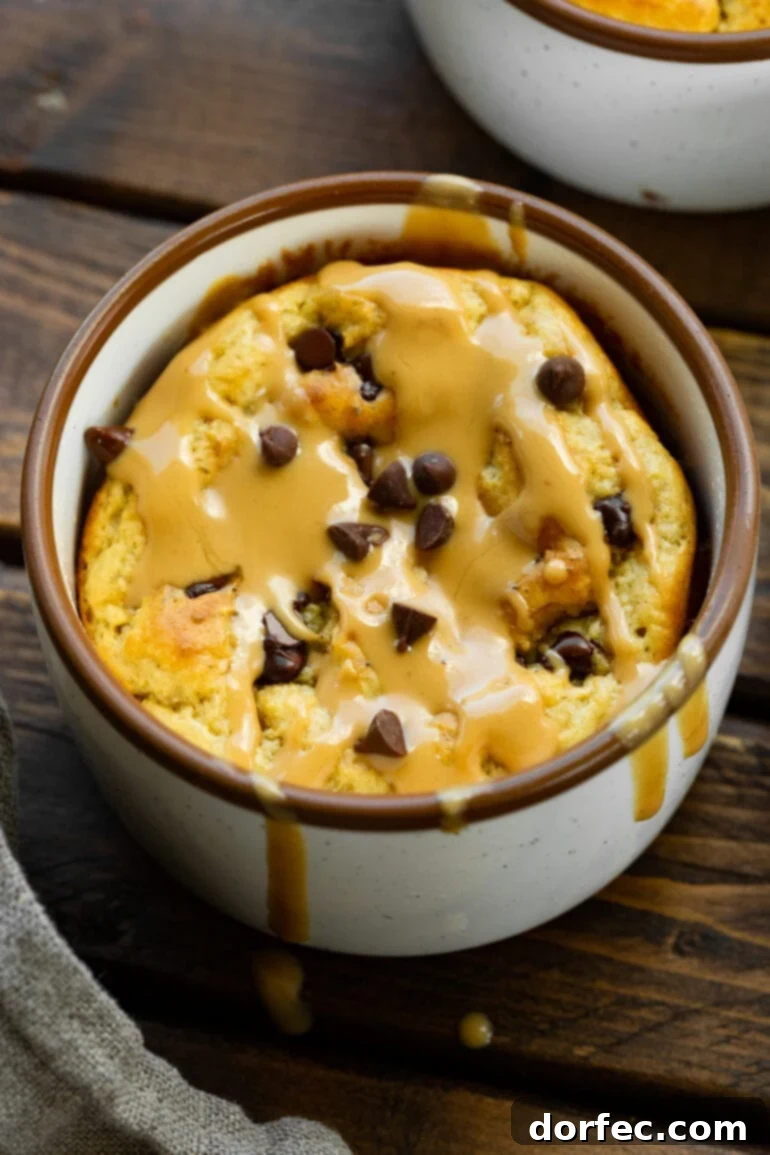 A close-up photo of a baked oatmeal mug cake, glistening with melted mini chocolate chips and a luscious drizzle of hot peanut butter.