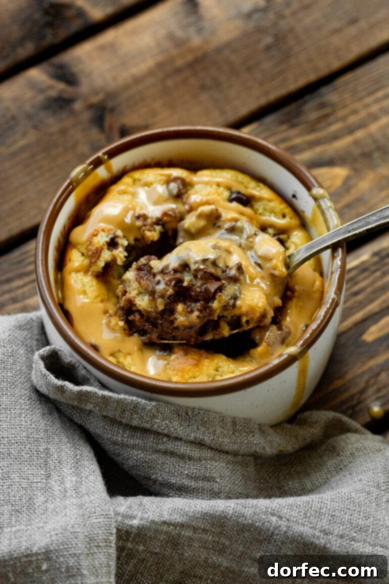 A hand scooping a generous portion of baked peanut butter chocolate chip oatmeal mug cake from a mug, showcasing its rich texture and topping of mini chocolate chips and melted peanut butter.