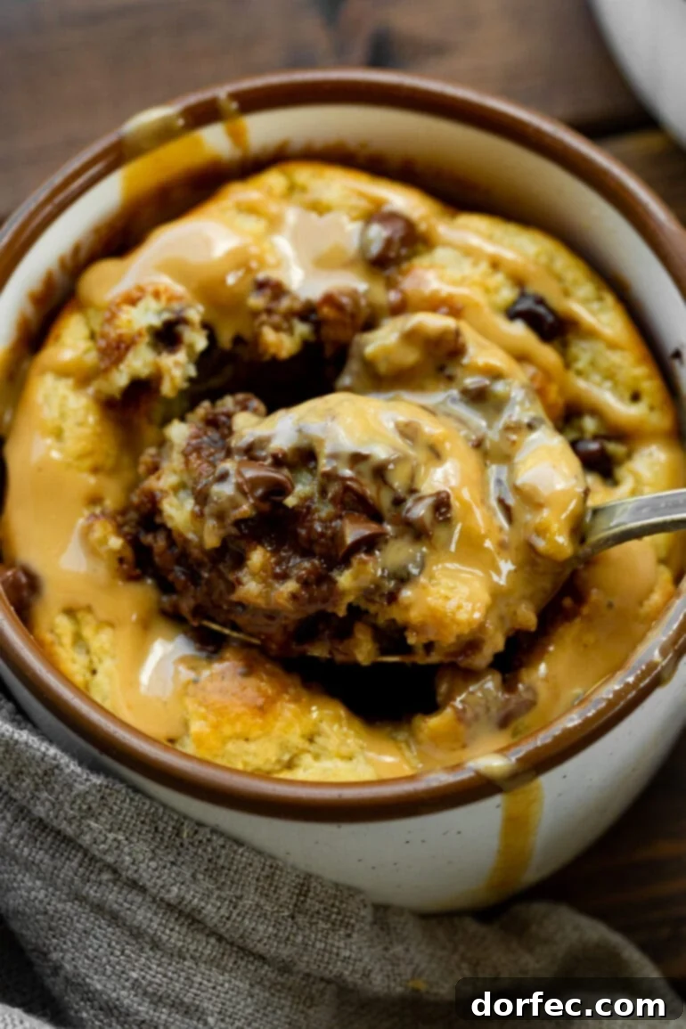A spoonful being carefully scooped out of the creamy center of a baked oatmeal mug cake, revealing melted chocolate chips and a rich peanut butter drizzle.