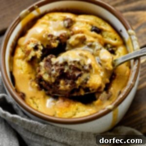 A photo of a mug full of baked oatmeal peanut butter and chocolate mug cake and someone is removing a spoonful of cake from the middle.