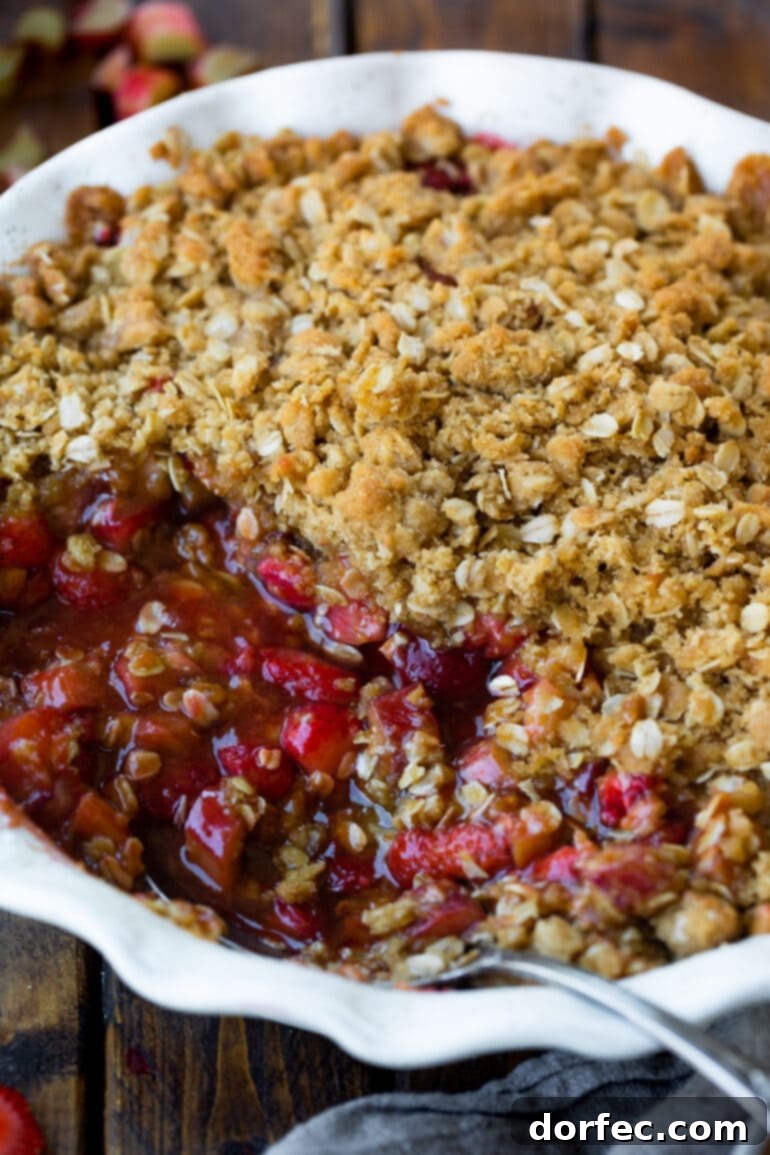 a photo of a strawberry rhubarb crisp in a circular white ceramic baking dish with a serving removed so you can see the juicy bright red fulling and butter oat topping.