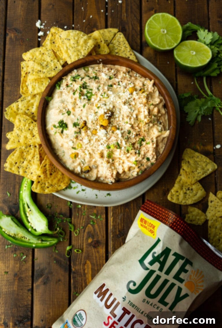 Zesty Mexican Street Corn Dip 4 a photo taken over the top of a large wooden bowl full of Mexican corn dip surrounded by tortilla chips and a bag of tortilla chips laying on its side and a halved jalapeno.