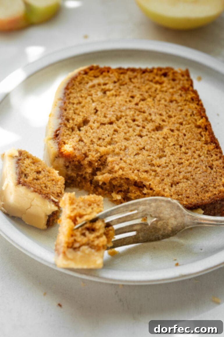 A tender slice of homemade apple bread with brown sugar icing rests on a white dessert plate, with a fork poised to take a bite, highlighting its moist texture.