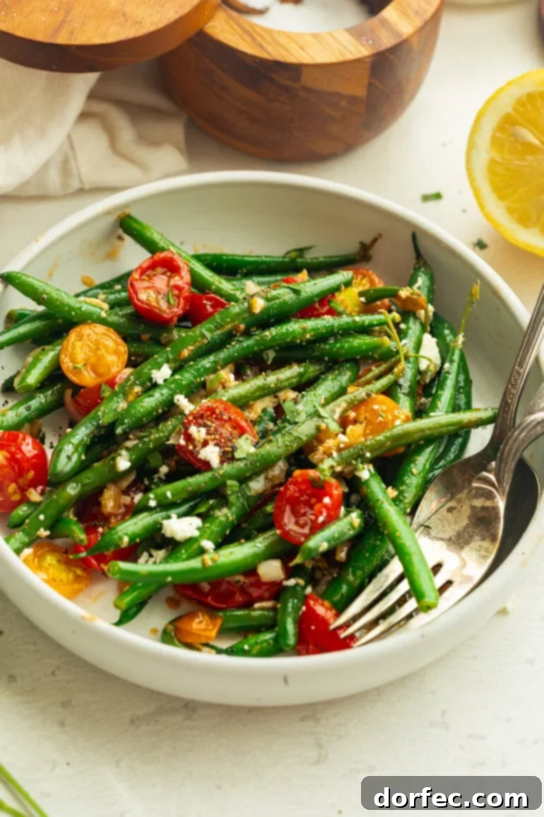 A close-up of vibrantly green sautéed green beans and cherry tomatoes in a white bowl, garnished with seasoning and a lemon wedge, ready to be served.