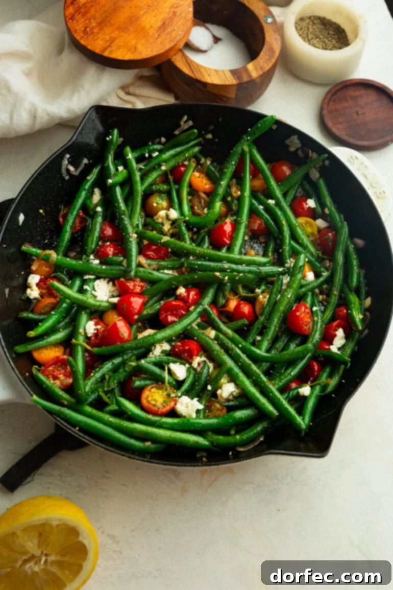 A cast iron skillet holds blistered cherry tomatoes and crisp-tender green beans, beautifully adorned with sliced garlic and fresh herbs.