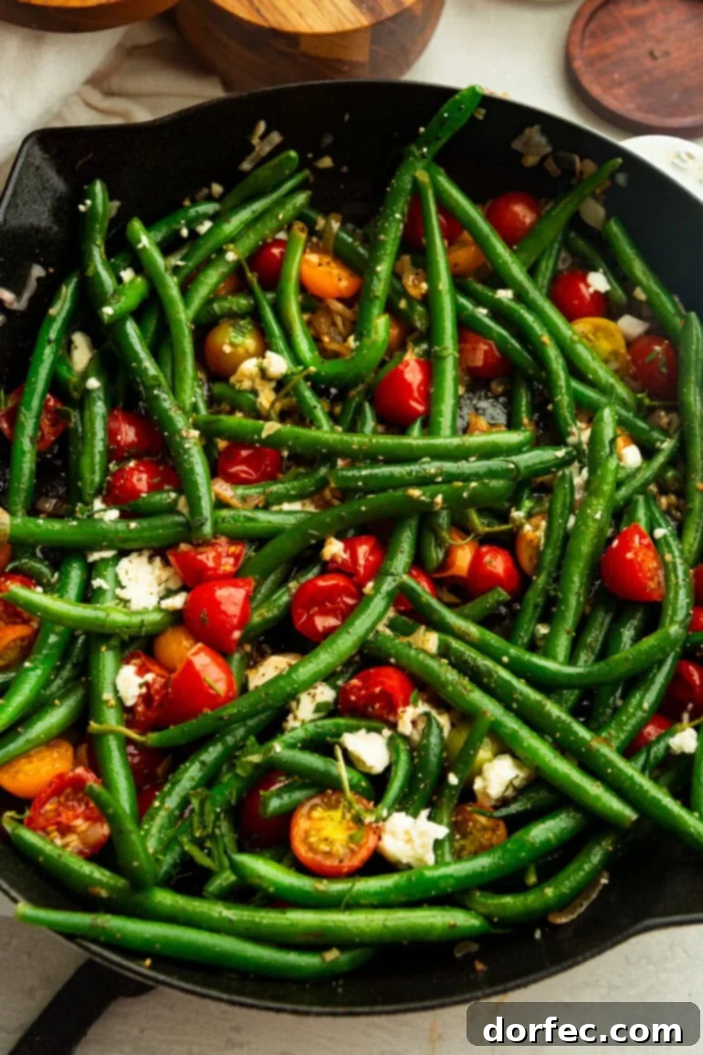 A top-down view of bright sautéed green beans and cherry tomatoes in a skillet, beautifully garnished with crumbled feta and freshly cracked black pepper.