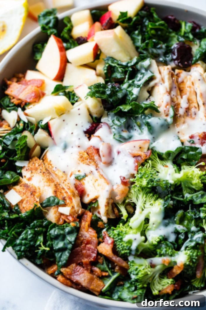A bountiful kale salad featuring crisp apples, savory bacon, dried cranberries, crunchy almonds, and slices of leftover smoked chicken, all coated in a poppy seed dressing.