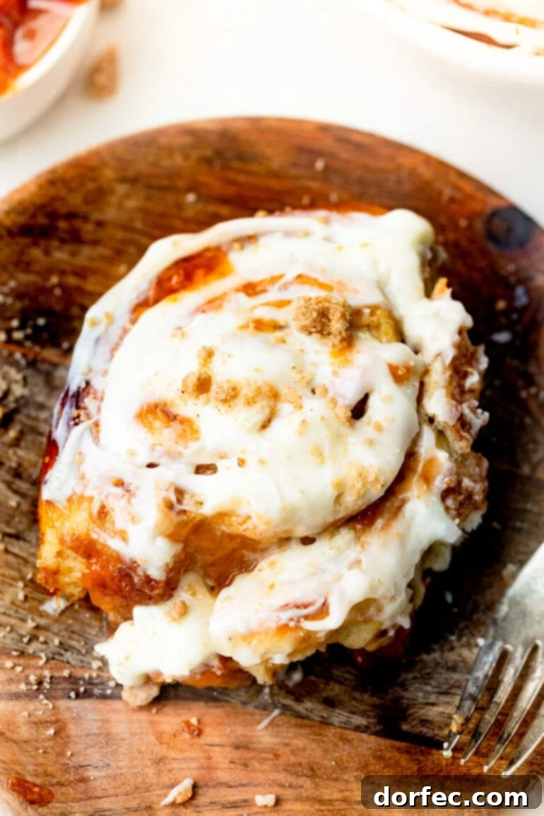 Peach Cobbler Swirl Buns 2 A close-up photo of a peach cinnamon roll on a wooden plate, overflowing with peach jam and generously topped with a rich peach cream cheese frosting, hinting at its ultimate deliciousness.