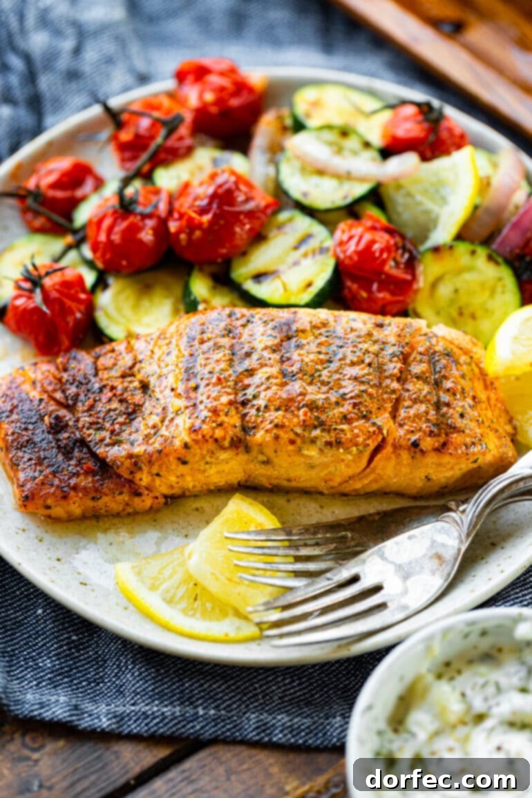 A perfectly grilled salmon fillet, flaked on one end to show its tender interior, is served on a dinner plate with a side of grilled zucchini and cherry tomatoes.