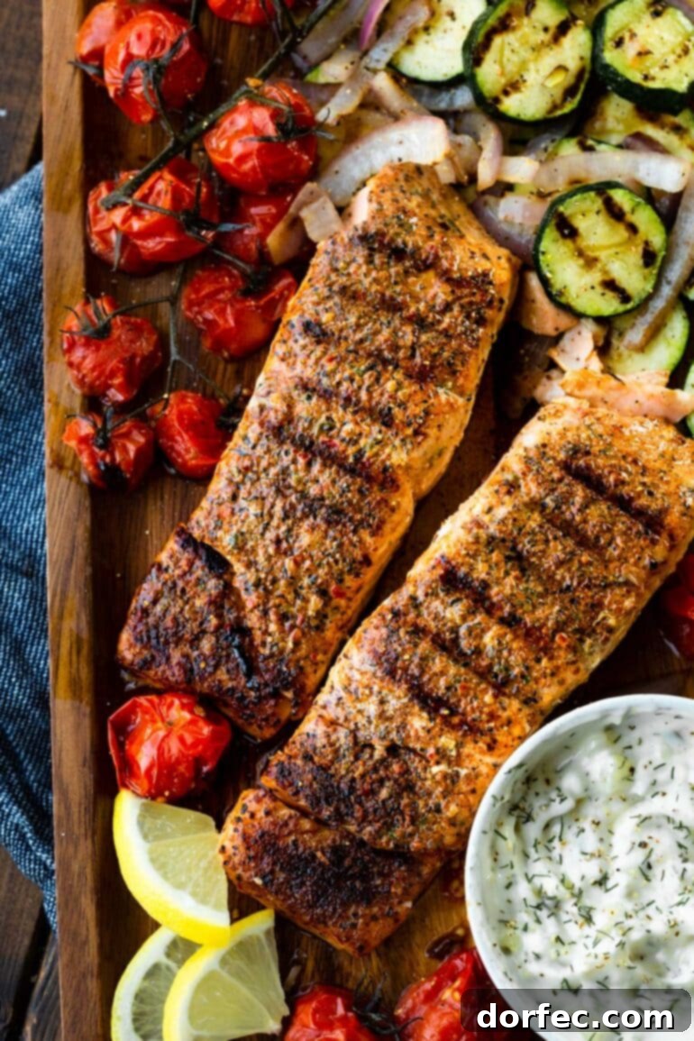 Two perfectly grilled salmon fillets are artfully arranged on a wooden serving tray, accompanied by a small bowl of creamy tartar sauce and a vibrant mix of grilled zucchini and cherry tomatoes.