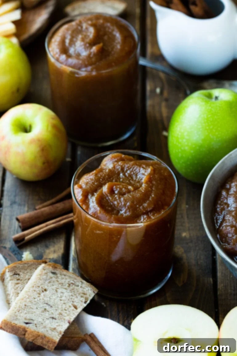 Set-It-and-Forget-It Slow Cooker Apple Butter 2 Two glasses full of rich, caramel-brown apple butter, surrounded by cinnamon sticks, whole apples, and peeled apple slices, evoking the warmth of autumn.