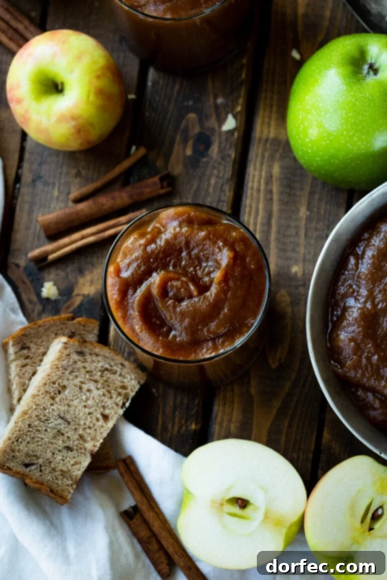 Set-It-and-Forget-It Slow Cooker Apple Butter 6 An overhead shot of a glass jar filled with luscious apple butter, artfully arranged with cinnamon sticks, whole and halved fresh apples, and a slice of rustic bread, highlighting its autumnal charm.