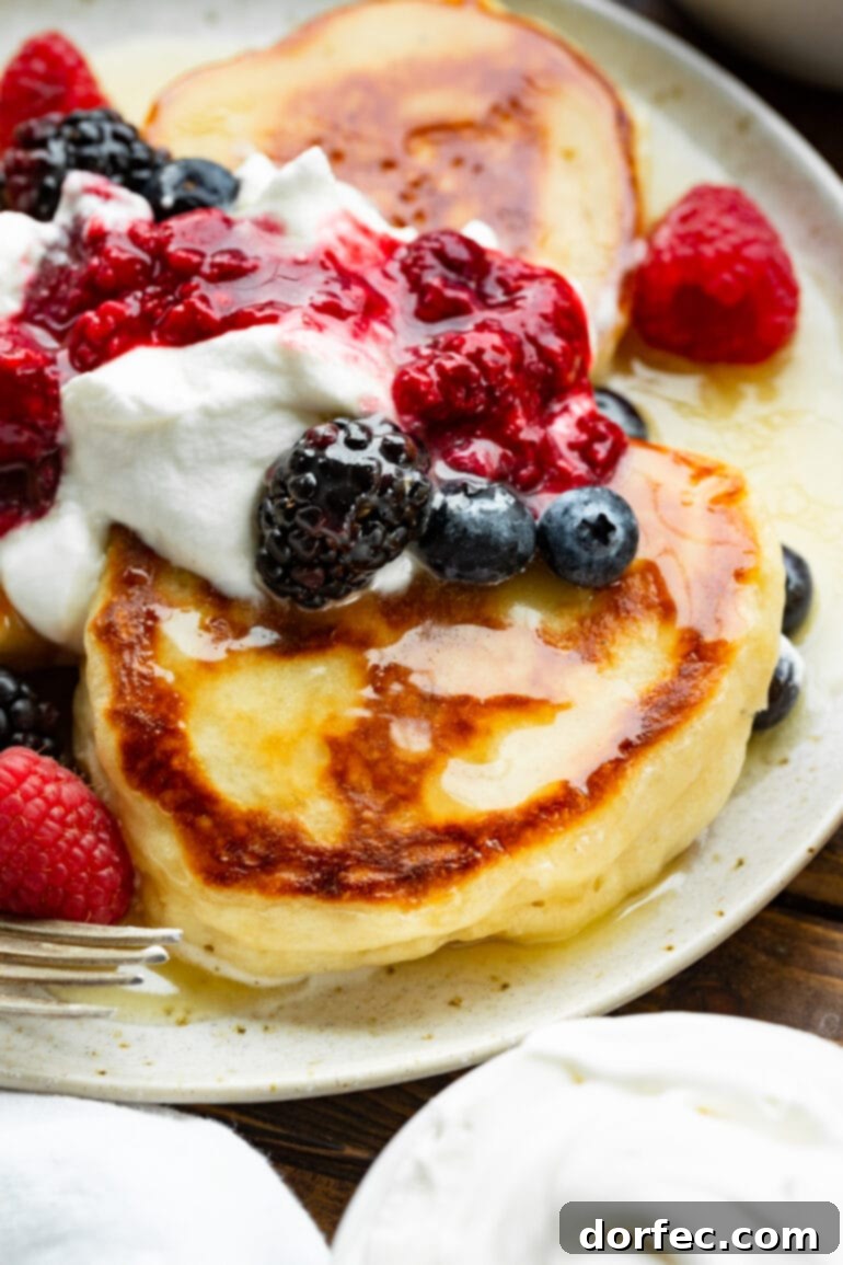 A close-up photograph of a single golden, crispy Oladi pancake, elegantly topped with a drizzle of syrup and a burst of fresh mixed berries.