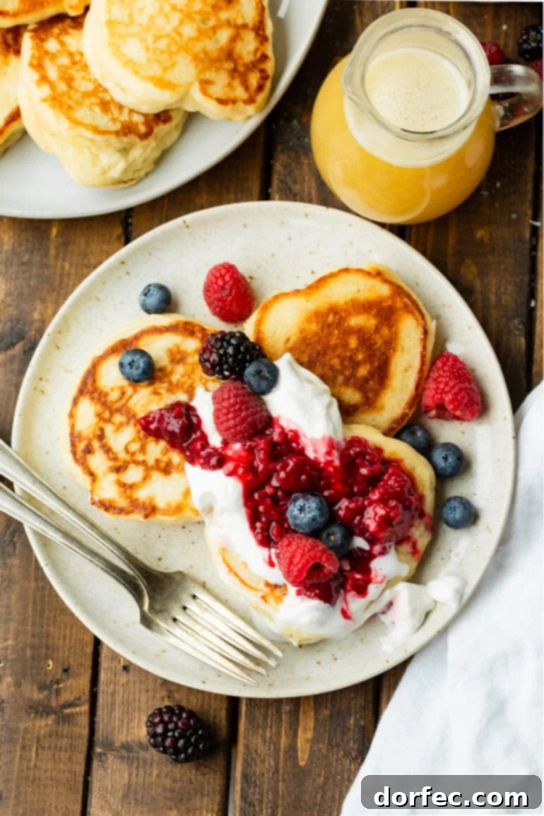 An overhead shot of a plate featuring three golden, crispy Oladi pancakes, artfully garnished with a medley of fresh mixed berries and a swirl of whipped cream.