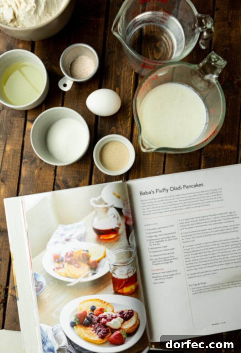 A beautifully composed photo showing an open cookbook displaying an Oladi pancake recipe, surrounded by individual small bowls and dishes containing all the precisely measured ingredients for the pancakes.