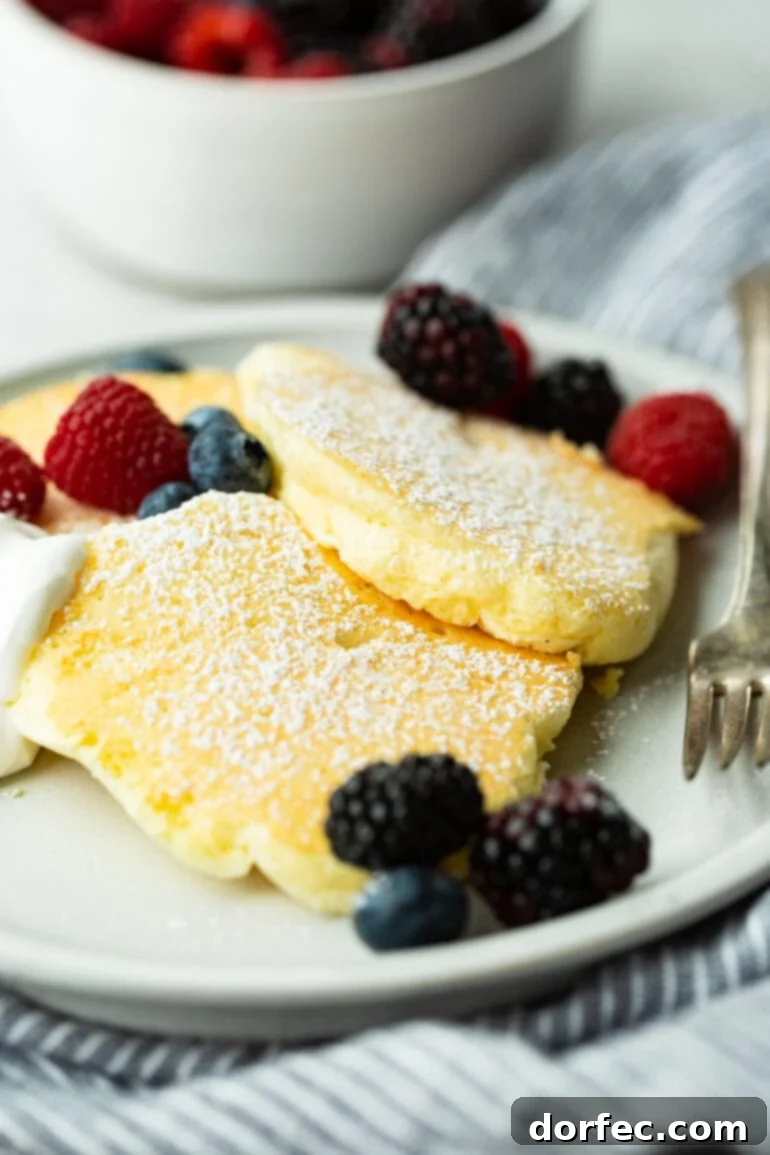 Fluffy Japanese Soufflé Pancakes 2 A mesmerizing photo of three exquisitely fluffy Japanese cloud pancakes, generously dusted with powdered sugar and adorned with a vibrant medley of fresh mixed berries. These golden-brown pancakes stand tall, showcasing their signature airy texture, a testament to their soufflé-like preparation.