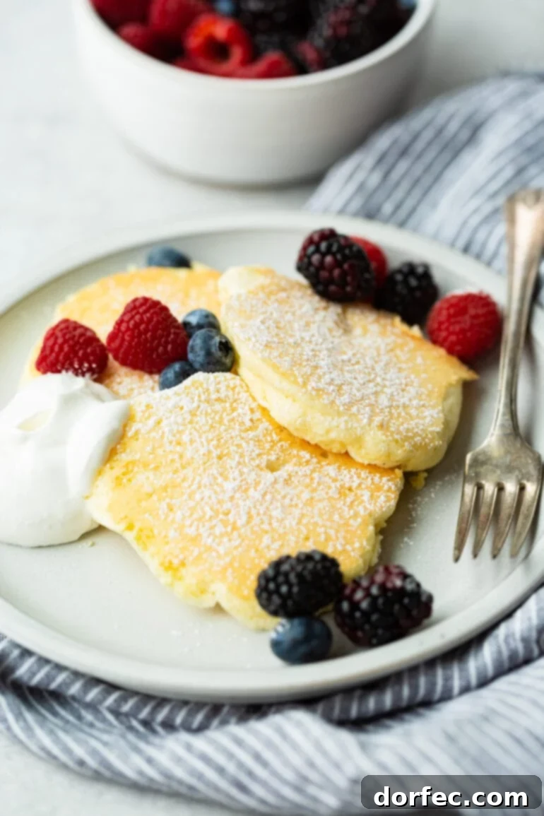 Fluffy Japanese Soufflé Pancakes 7 A beautifully composed photo of three pristine Japanese cloud pancakes resting on a white plate. They are elegantly topped with a dusting of powdered sugar, encircled by a scattering of fresh mixed berries, and accompanied by a dollop of luscious whipped cream on the side, creating a delightful and inviting presentation.