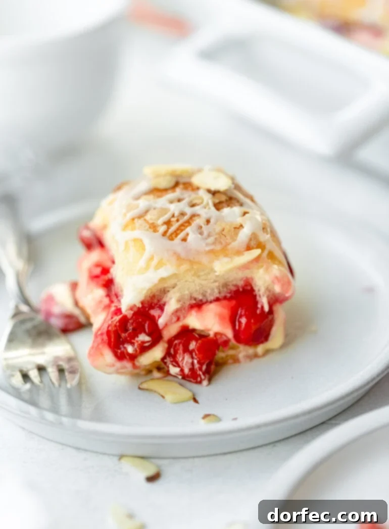 A close-up photo of a golden-baked cherry cheese danish slider, topped with a sweet white glaze and sliced almonds, with a silver fork resting beside it, inviting a bite.