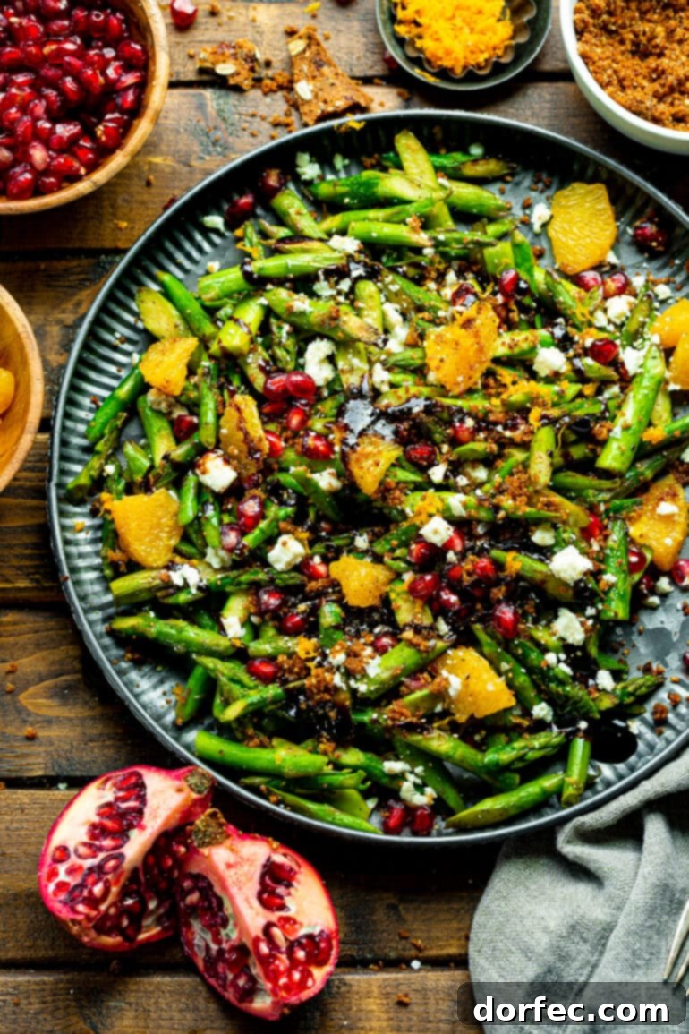 A beautifully arranged serving platter showcasing a festive asparagus salad, adorned with jewel-like pomegranate arils, bright orange segments, crumbles of Gorgonzola cheese, and a elegant drizzle of balsamic glaze.