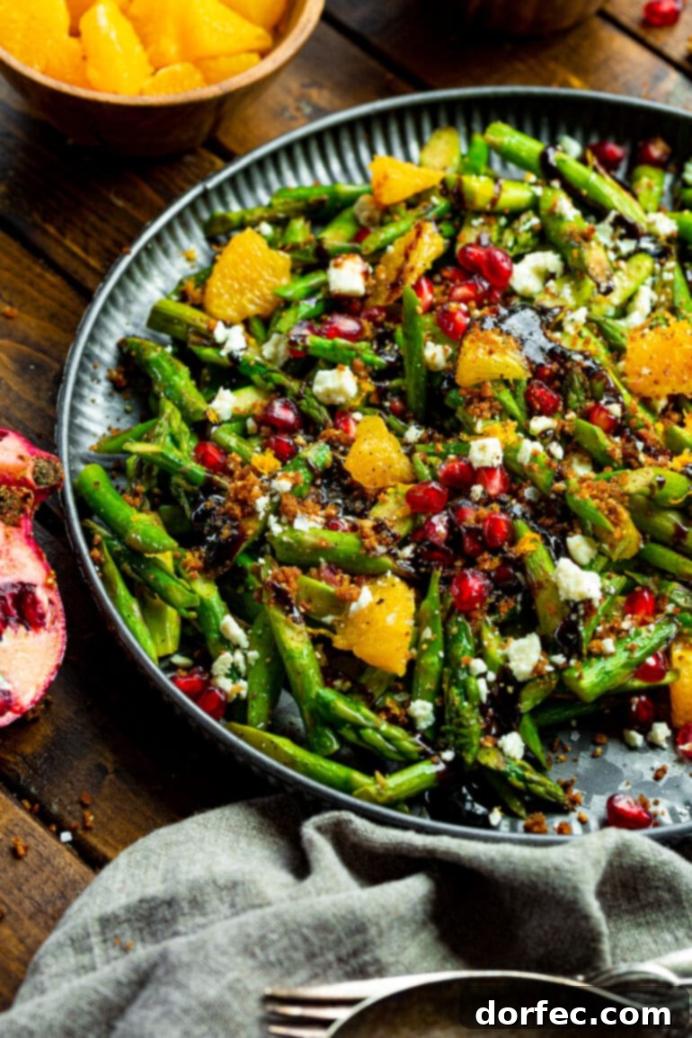 A large, elegant serving tray brimming with a holiday asparagus salad, featuring a generous mix of bright green asparagus, ruby red pomegranate seeds, sweet orange wedges, creamy Gorgonzola cheese, toasted panko, and a glistening balsamic glaze.