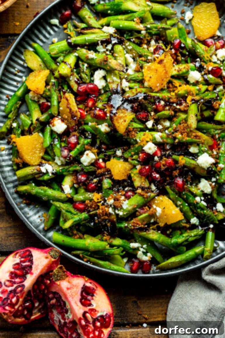 A large, brightly lit serving tray overflowing with a fresh, colorful asparagus, pomegranate, and orange salad, garnished with crumbled cheese and a balsamic drizzle.