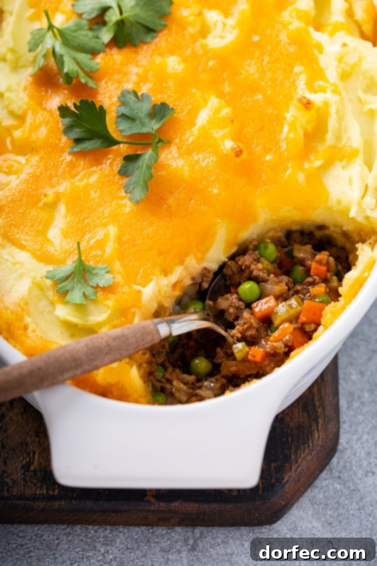 A casserole dish containing Shepherd's Pie with a spoonful removed, revealing layers of hamburger, carrots, and peas topped with mashed potatoes and melted cheddar cheese. A classic comfort food dish.