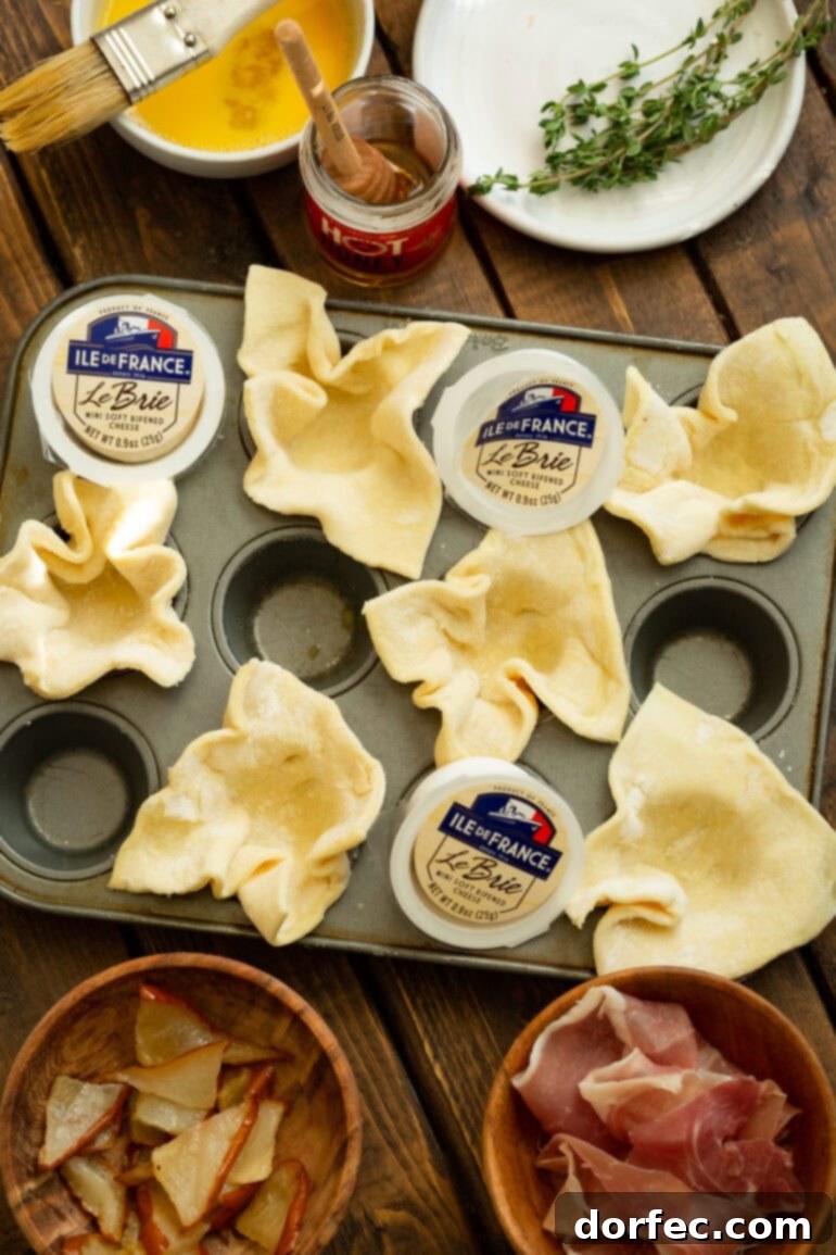 A photo of a small muffin tin with half the cups containing unbaked puff pastry and small pieces of Brie in other cups, showcasing the assembly process.