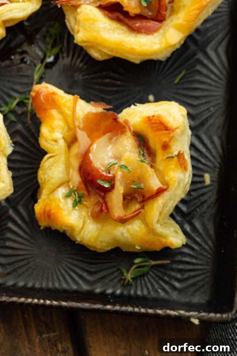 A photo taken over the top of a golden-baked puff pastry bite full of roasted pear, brie, prosciutto, and fresh thyme, beautifully garnished.