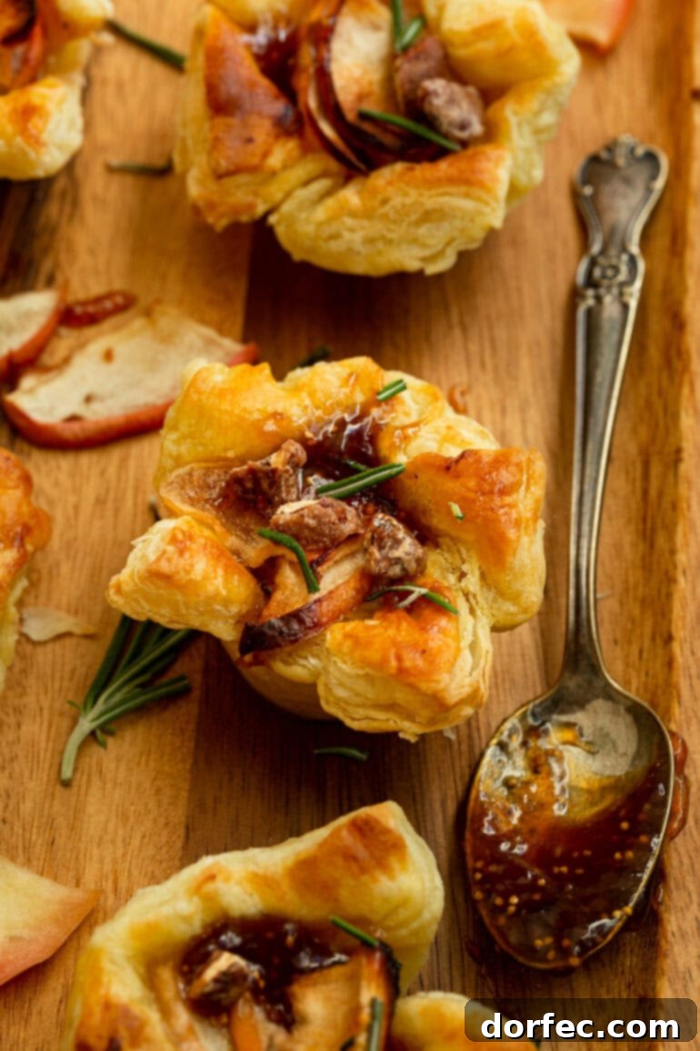 a photo of a golden baked puff pastry appetizer topped with roasted apples, fresh rosemary, candied pecans with a spoonful of fig jam on the side