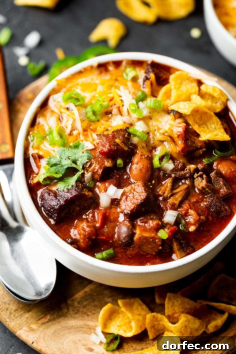 A close-up photo of a white soup bowl filled with homemade brisket chili, topped with Fritos, melting shredded cheese, and green onions.