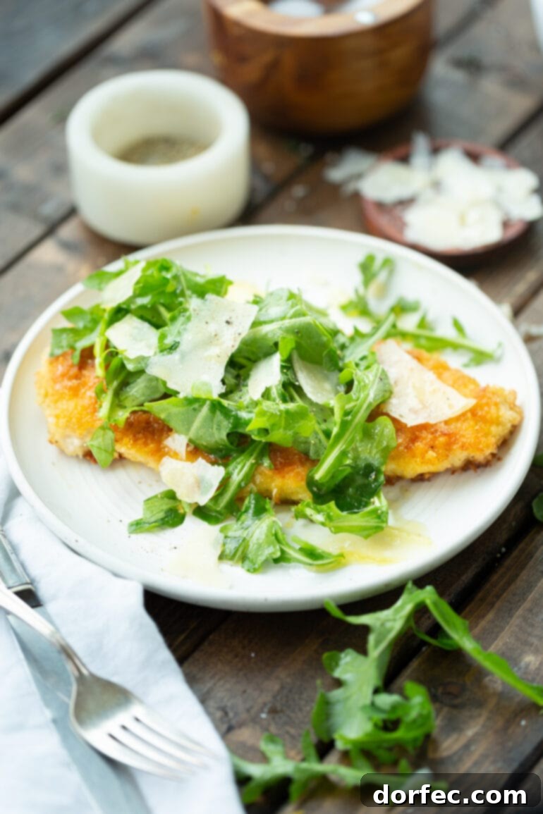 A golden breaded chicken breast, lightened up, served on a white dinner plate, topped with fresh salad greens and shaved Parmesan cheese, with a lemon vinaigrette.