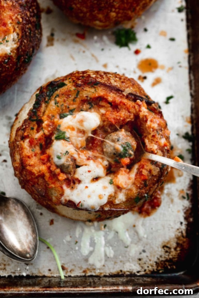 A rustic bread bowl generously filled with bubbling Parmesan Meatball Soup, topped with melted cheese and a spoon ready for enjoyment. A perfect cozy meal for chilly weather.