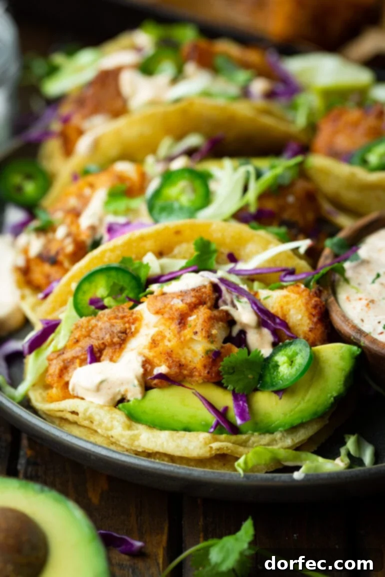 A vibrant tray full of golden-brown fish tacos, generously topped with shredded purple and green cabbage, thinly sliced jalapeños, creamy avocado slices, and a drizzle of rich, white fish taco sauce.