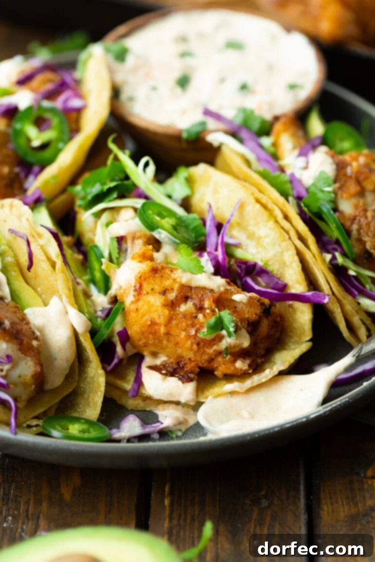 A striking photo of several homemade fish tacos neatly arranged on a plate, adorned with vibrant shredded green and purple cabbage, glistening avocado and jalapeño slices, and a generous drizzle of the creamy white fish taco sauce.