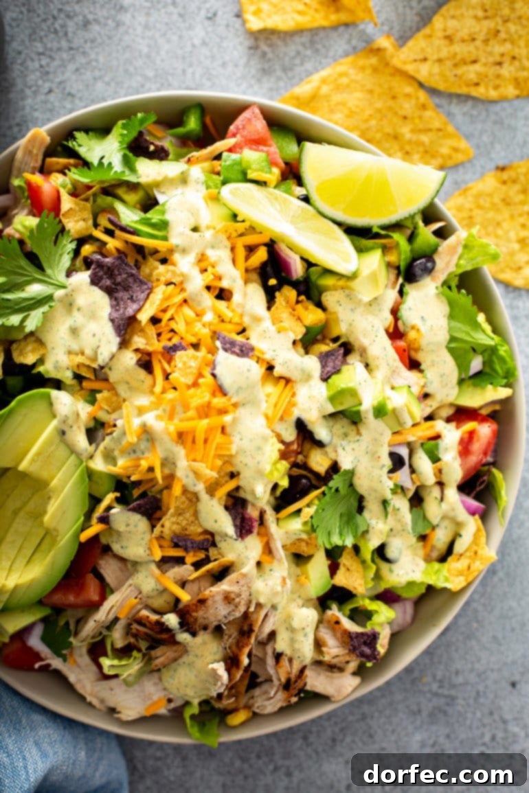 A large, colorful bowl of chicken taco salad, featuring shredded chicken, cheese, cilantro, black beans, corn, diced tomatoes, red onions, bell peppers, and sliced avocados. Two lime wedges, tortilla chips, and a bottle of dressing are visible in the background.