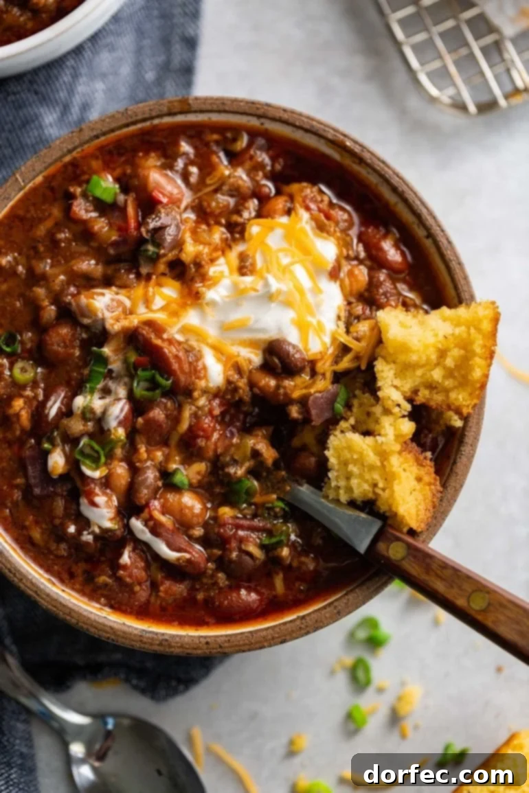 A steaming bowl of rich, homemade chili, featuring ground beef, multiple types of beans, and a flavorful sauce. It's generously topped with sour cream and shredded cheese, with a golden piece of cornbread resting on the rim of the bowl.