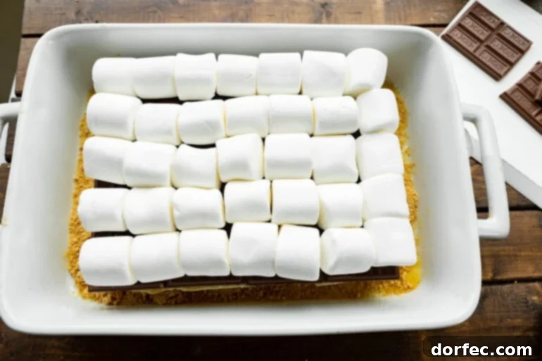 S'mores Sliders 5 The process of assembling s'mores sliders, showing the bottom half of a dozen slider buns topped with layers of Hershey's chocolate bars and large marshmallows, ready for the top bun.