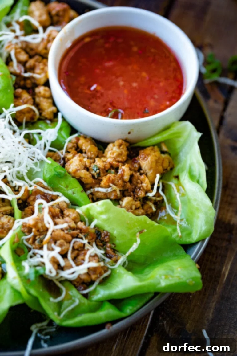 A serving of flavorful Asian chicken lettuce wraps, beautifully arranged and topped with crispy rice noodles, accompanied by a small bowl of sweet chili dipping sauce.