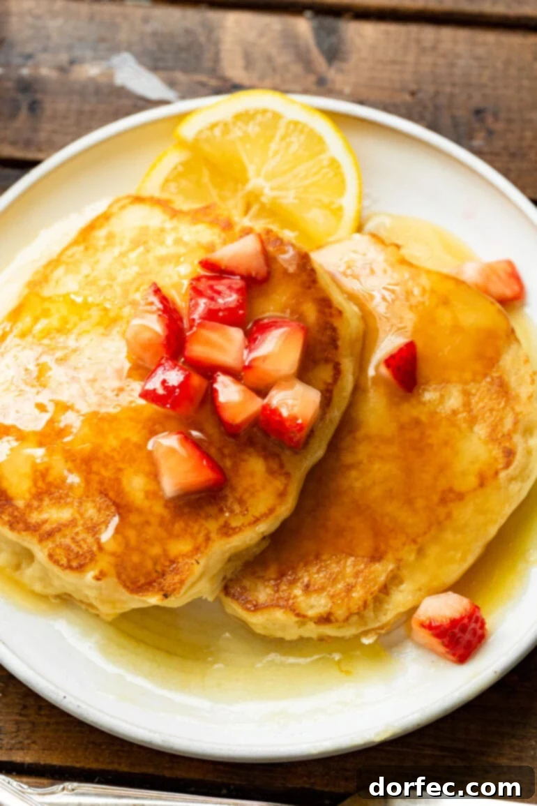 A stack of perfectly golden and fluffy buttermilk pancakes, generously topped with fresh diced strawberries and drizzled with luscious buttermilk syrup, served on a white plate.