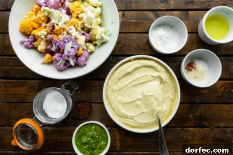 A vibrant flat lay photo showcasing all the fresh ingredients required for making crispy roasted cauliflower and hummus, including a bowl of creamy hummus, a small dish of bright green pesto sauce, and a colorful plate of orange and purple cauliflower florets.